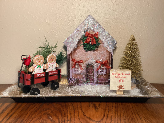 Adorable table top snow-covered gingerbread house with gingerbread cookies in a wagon, gold Christmas tree